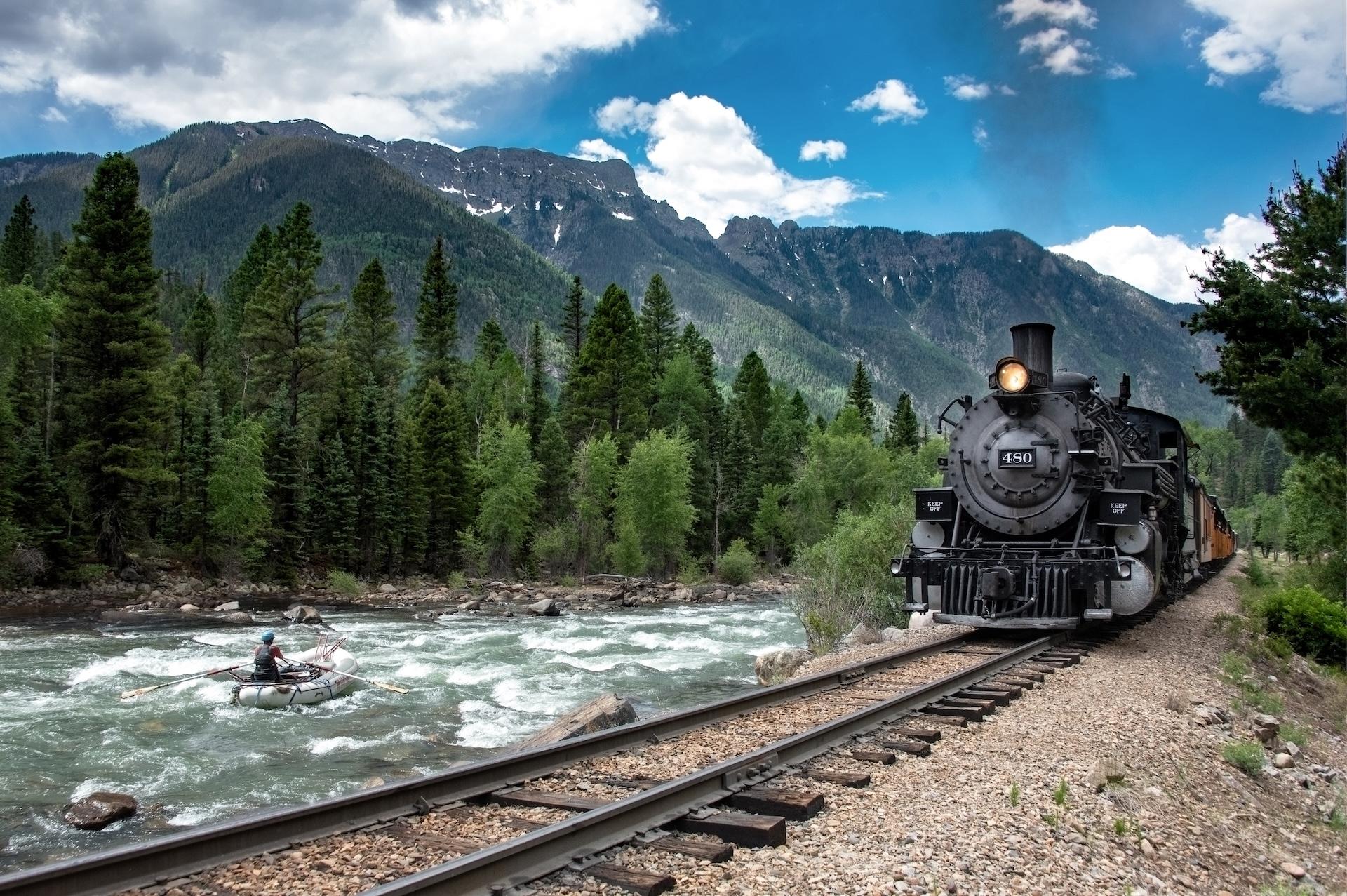 Train traveling alongside a river with mountains in the background